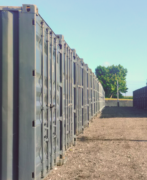 Long rang de boxes de stockage en acier gris, alignés sur un terrain gravillonneux sous un ciel bleu.